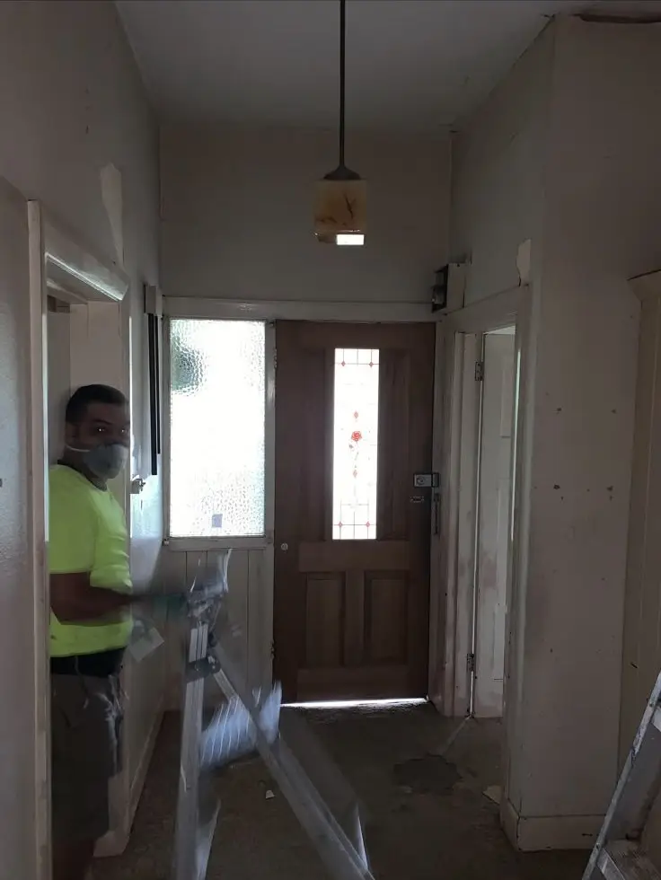 Worker in a neon shirt and mask stands in a hallway with protective plastic sheets and a wooden front door.