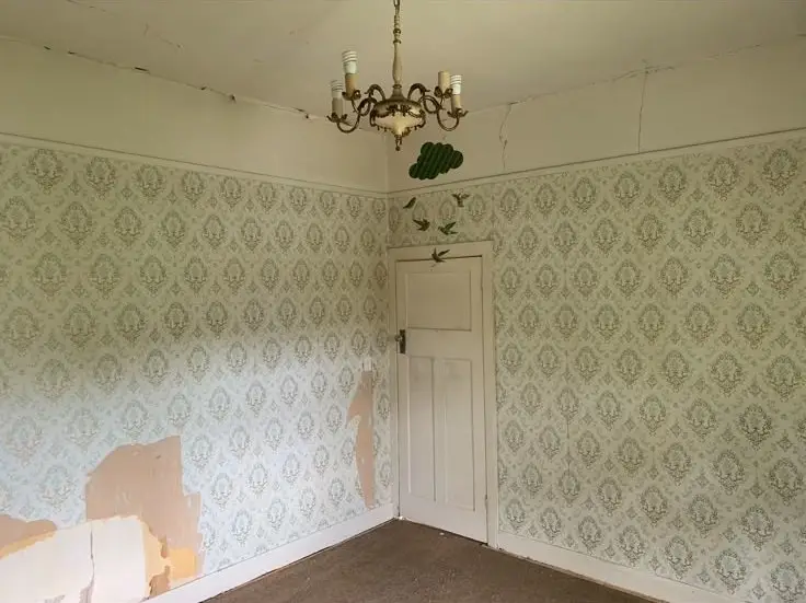 Old empty room with ornate wallpaper, peeling patches on the left wall, a vintage chandelier, and a closed white door.