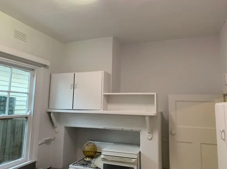 White kitchen area with a stove beneath a mantel-like shelf, a wall cabinet, and an open shelf above; window on the left lets in light.