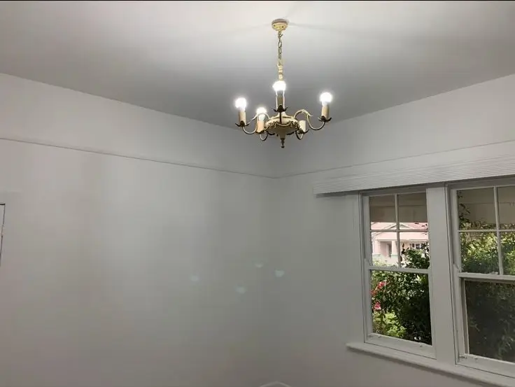 Bright white room corner with a brass chandelier hanging from the ceiling and a large window showing greenery outside.