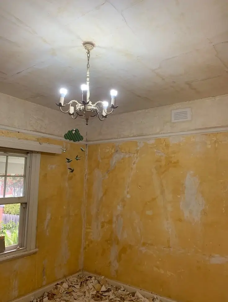Room under renovation with yellow plaster walls, a damaged ceiling, and debris on the floor; a chandelier hangs from the ceiling near a window.
