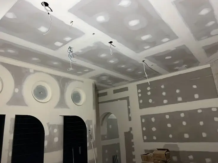 Room under drywall installation with taped seams, hanging electrical wires from the ceiling, circular wall vents, and an arched doorway.
