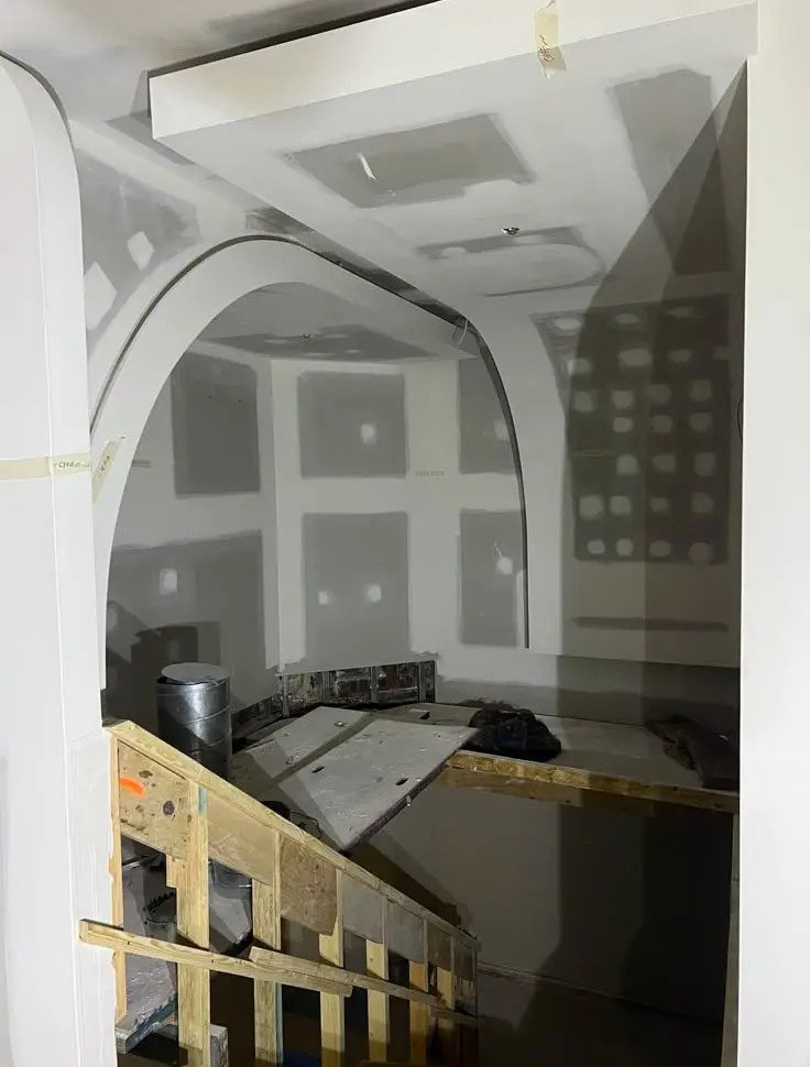 Under-construction interior featuring a curved white archway and drywall panels on the walls, with wooden scaffolding and a work platform in the foreground.