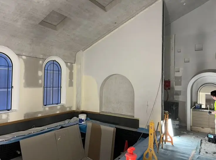 Interior under renovation: arched blue windows, partially finished walls, and construction materials scattered along a long counter area.