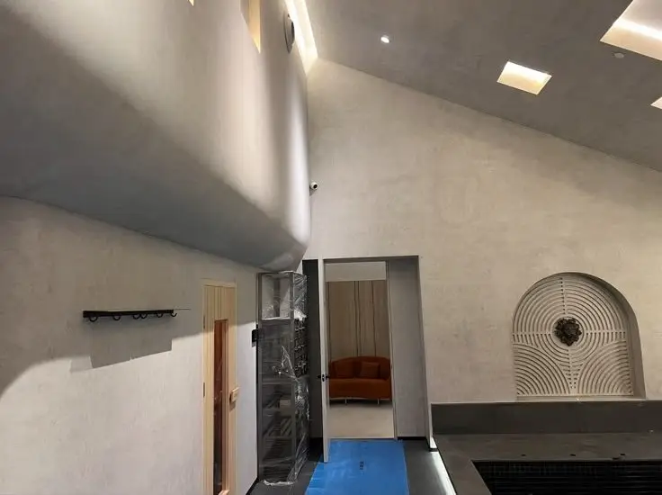 Modern, minimalist interior with concrete walls and curved upper balcony; open doorway leads to a room with an orange sofa.